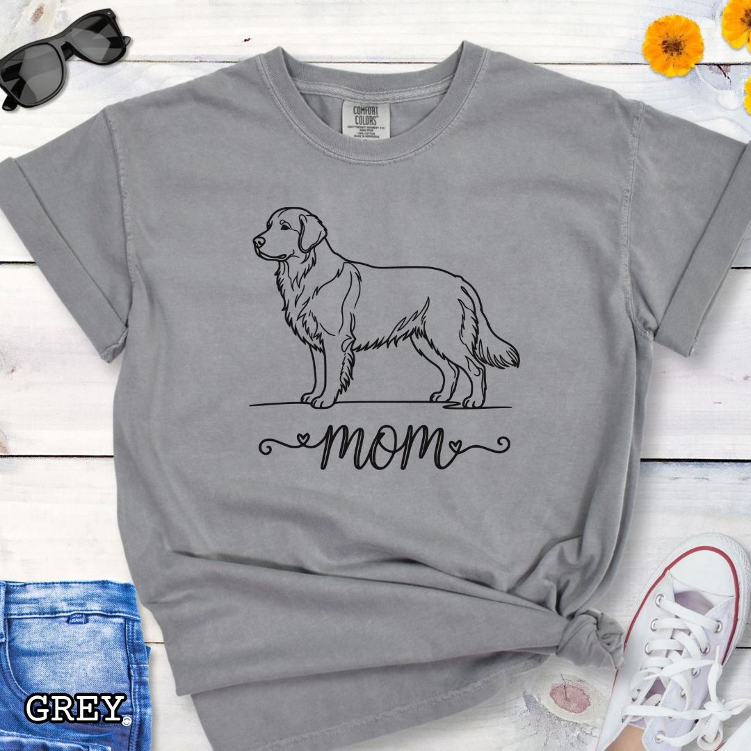 Grey Comfort Colors t-shirt with a line art Golden Retriever dog illustration and 'mom' text in feminine script beneath, shown on a wooden surface with denim jeans and a flower.