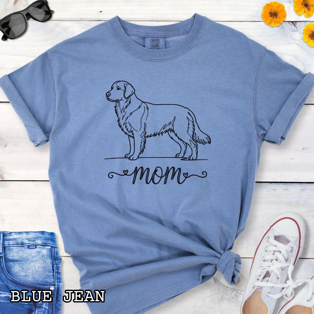 Blue Comfort Colors t-shirt with a line art Golden Retriever dog illustration and 'mom' text in feminine script beneath, shown on a wooden surface with denim jeans and a flower.