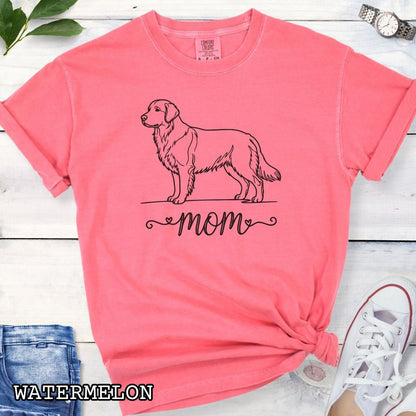 Pink Comfort Colors t-shirt with a line art Golden Retriever dog illustration and 'mom' text in feminine script beneath, shown on a wooden surface with denim jeans and a flower.