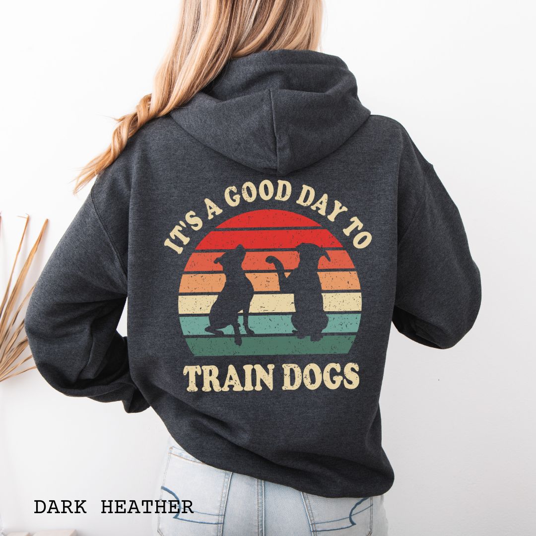 Dark grey back print hoodie with retro sunset and silhouettes of two sitting dogs, and the text It's A Good Day to Train Dogs, presented on a wooden surface with jeans and shoes.