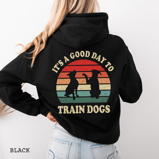 Black back print hoodie with retro sunset and silhouettes of two sitting dogs, and the text It's A Good Day to Train Dogs, presented on a wooden surface with jeans and shoes.
