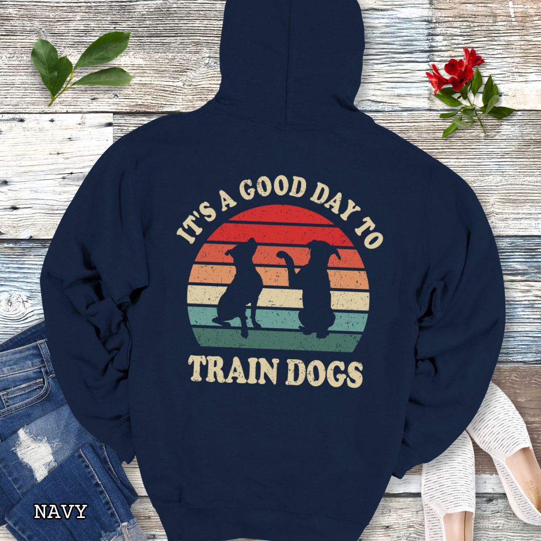 Navy back print hoodie with retro sunset and silhouettes of two sitting dogs, and the text It's A Good Day to Train Dogs, presented on a wooden surface with jeans and shoes.