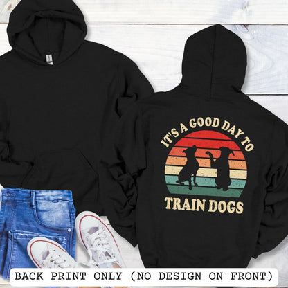 Black back print hoodie with retro sunset and silhouettes of two sitting dogs, and the text It's A Good Day to Train Dogs, presented on a wooden surface with jeans and shoes.