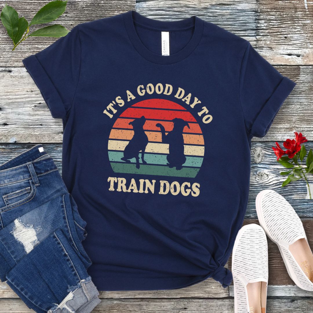 Navy blue t-shirt with retro sunset and silhouettes of two sitting dogs, and the text It's A Good Day to Train Dogs, presented on a wooden surface with jeans and shoes.