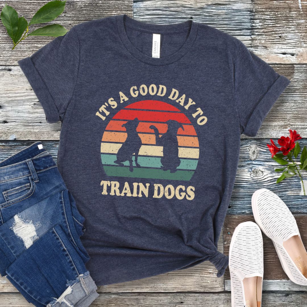 Heather navy t-shirt with retro sunset and silhouettes of two sitting dogs, and the text It's A Good Day to Train Dogs, presented on a wooden surface with jeans and shoes.