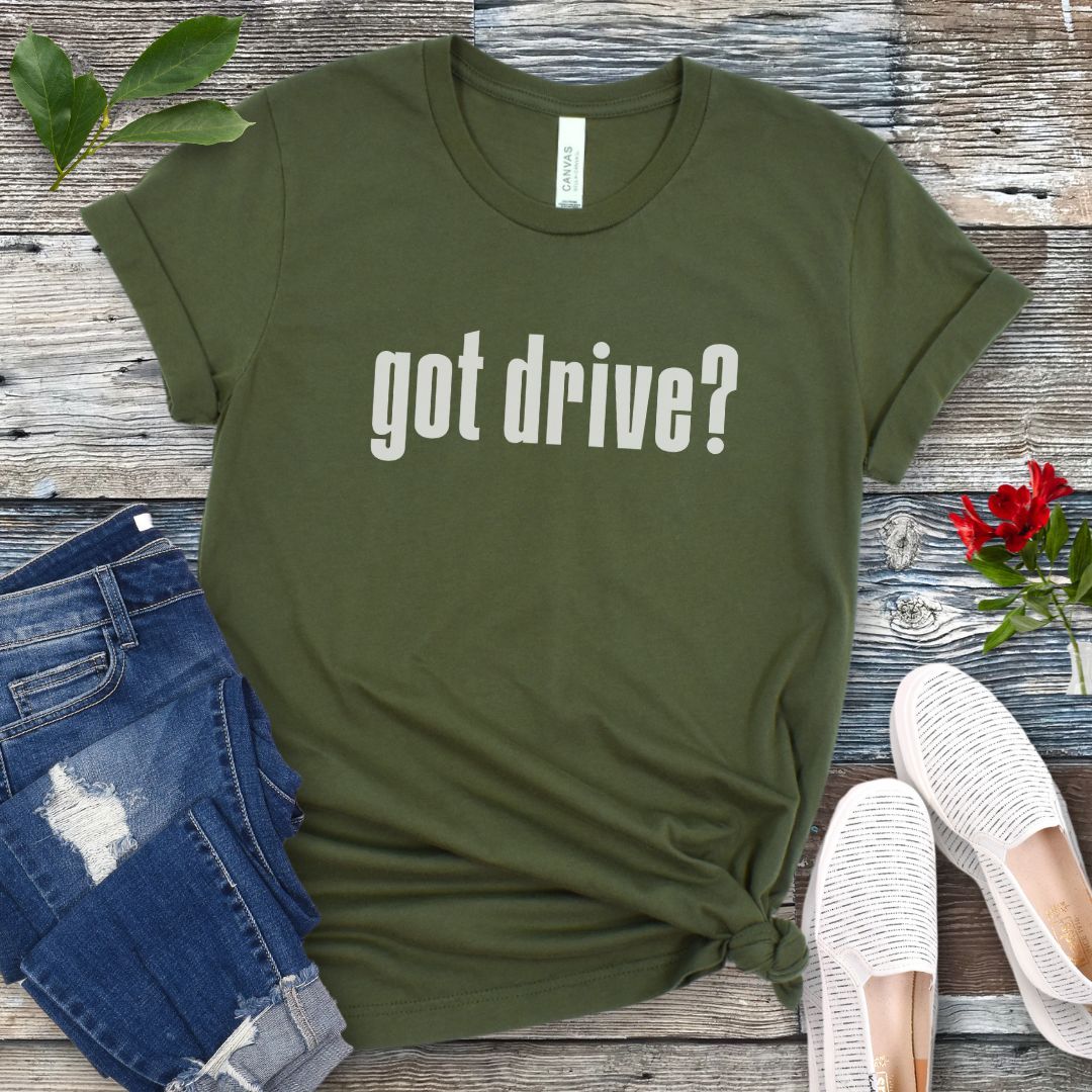 Military green IGP dog sports t-shirt with 'got drive?' text on a wooden surface with jeans and shoes.