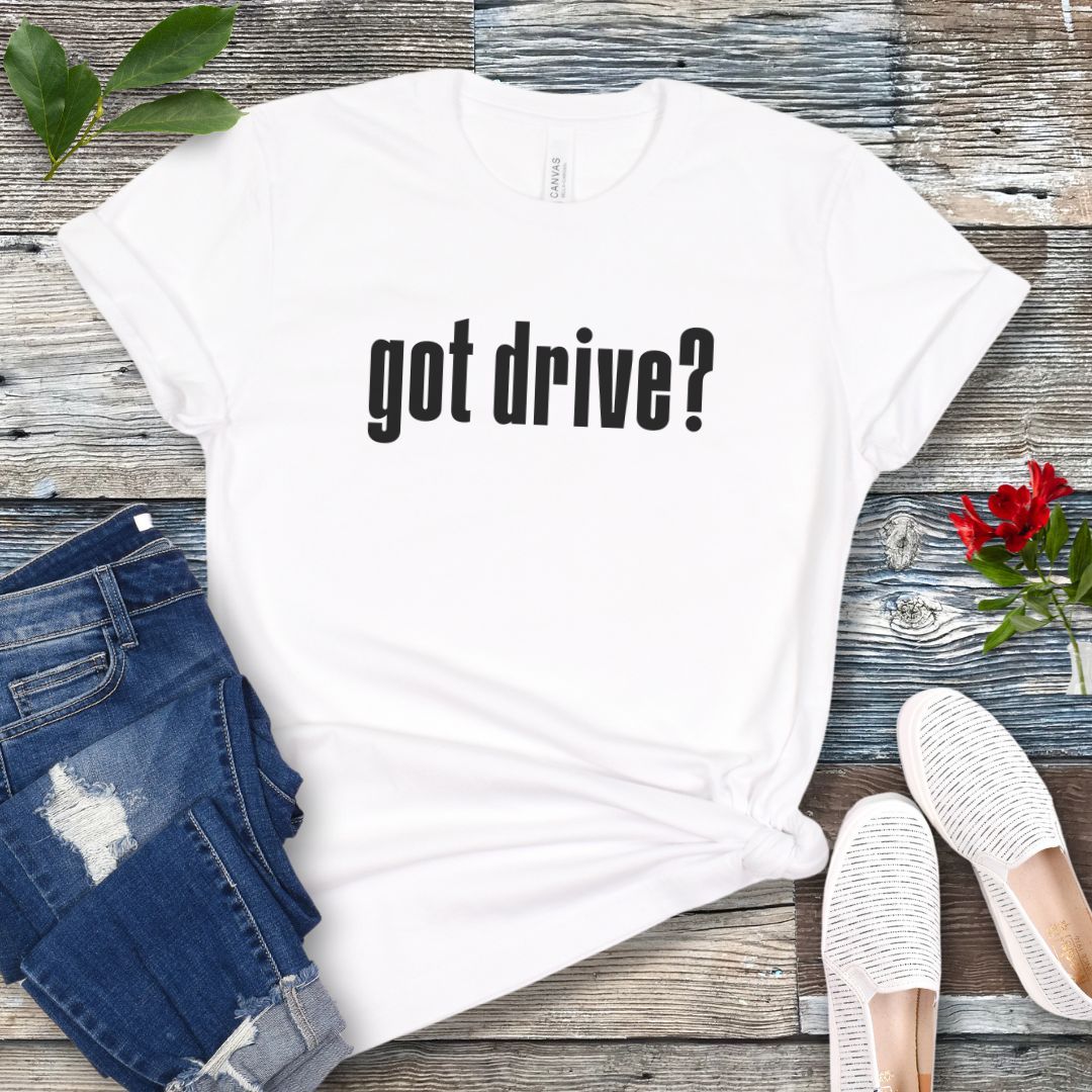 White IGP dog sports t-shirt with 'got drive?' text on a wooden surface with jeans and shoes.