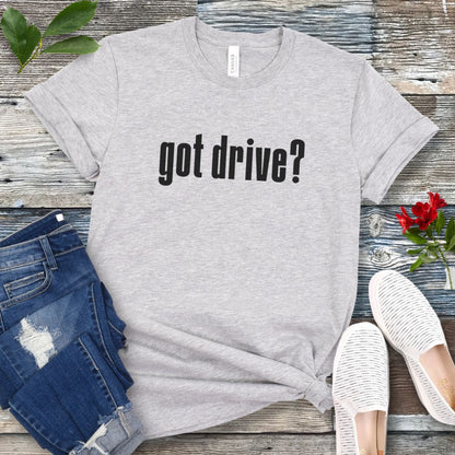 Grey IGP dog sports t-shirt with 'got drive?' text on a wooden surface with jeans and shoes.