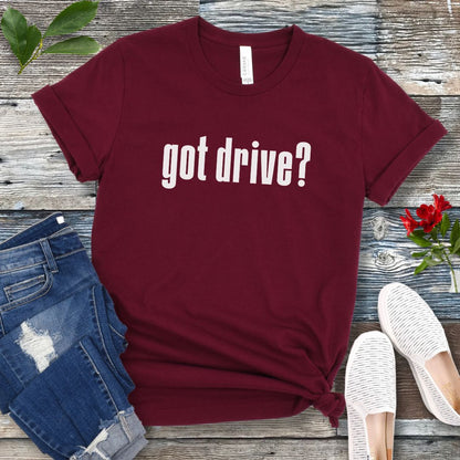Maroon IGP dog sports t-shirt with 'got drive?' text on a wooden surface with jeans and shoes.