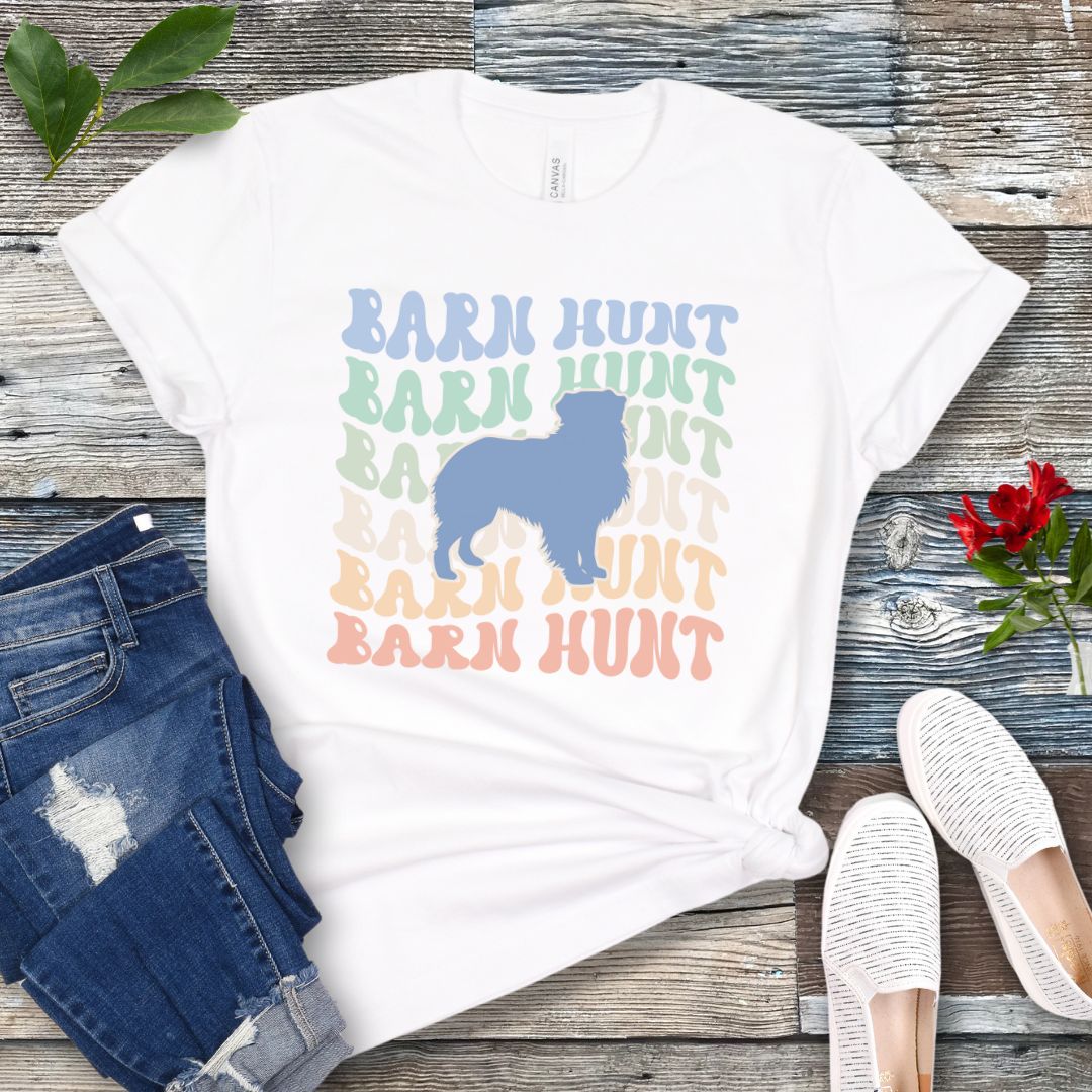 White dog sports t-shirt with 'Barn Hunt' repeated in colorful retro style text and a custom dog breed silhouette, presented on a wooden surface with jeans and shoes.