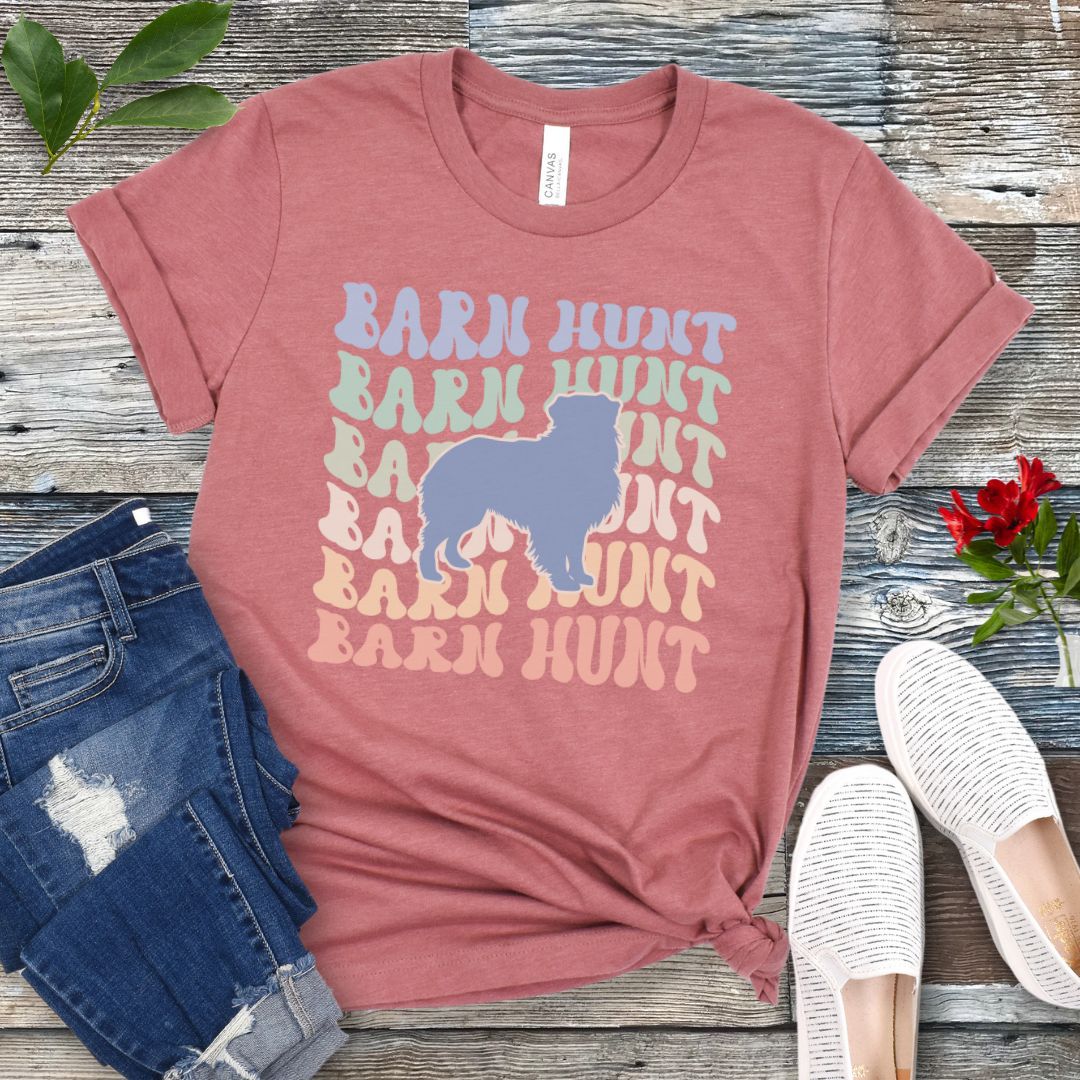Mauve dog sports t-shirt with 'Barn Hunt' repeated in colorful retro style text and a custom dog breed silhouette, presented on a wooden surface with jeans and shoes.