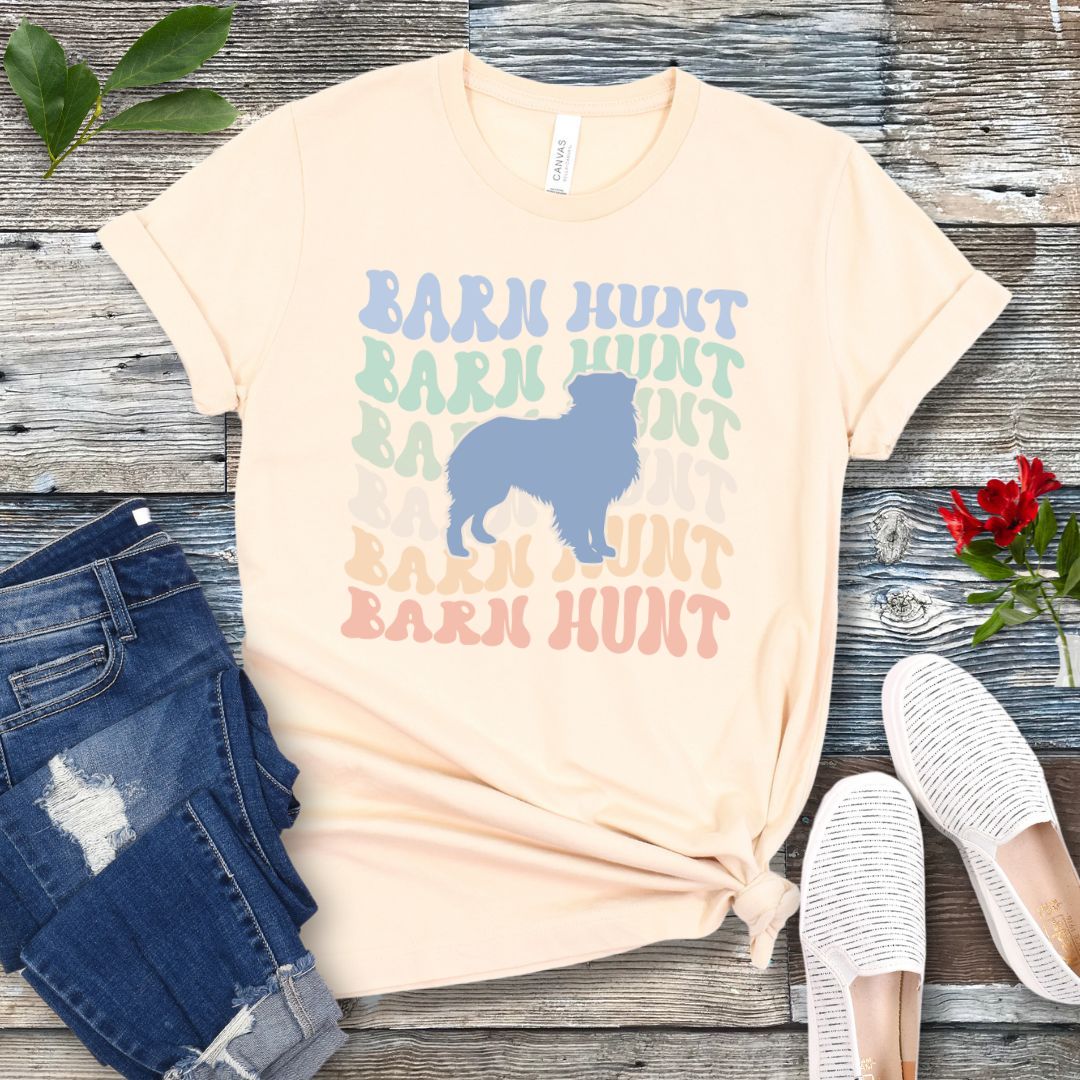 Beige dog sports t-shirt with 'Barn Hunt' repeated in colorful retro style text and a custom dog breed silhouette, presented on a wooden surface with jeans and shoes.