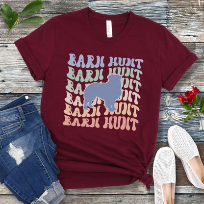 Maroon dog sports t-shirt with 'Barn Hunt' repeated in colorful retro style text and a custom dog breed silhouette, presented on a wooden surface with jeans and shoes.