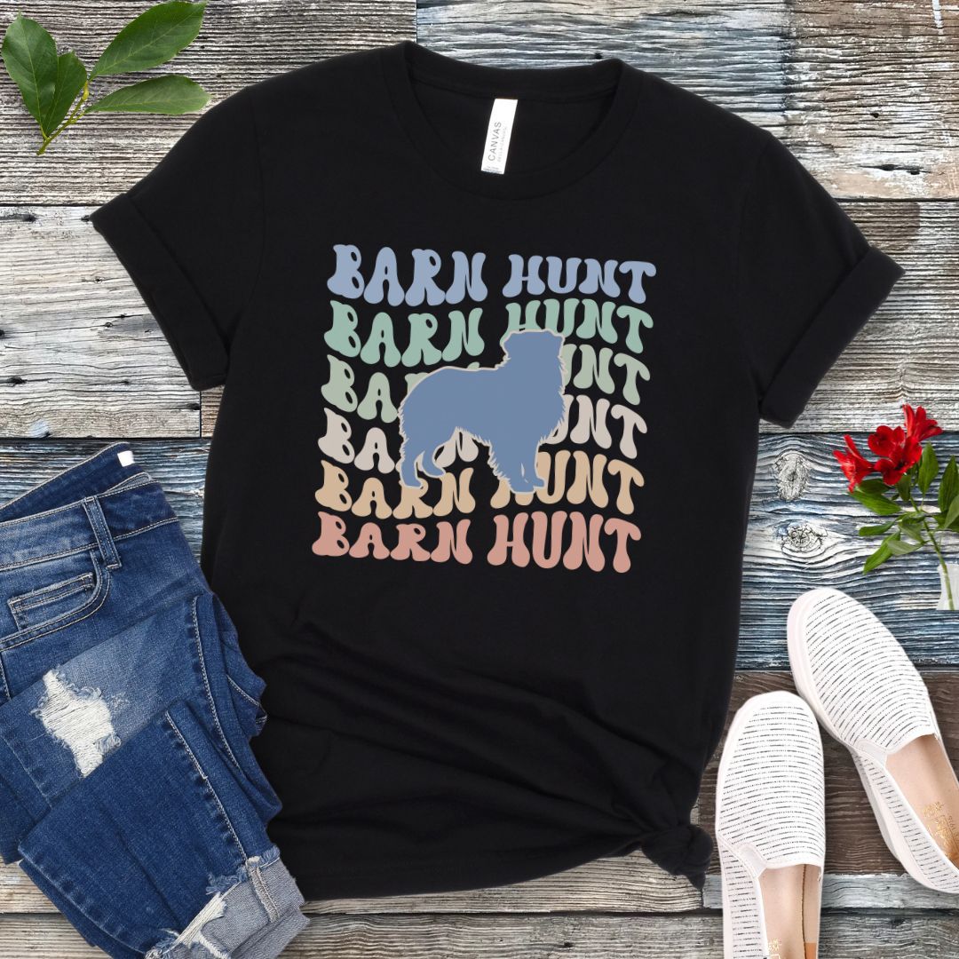 Black dog sports t-shirt with 'Barn Hunt' repeated in colorful retro style text and a custom dog breed silhouette, presented on a wooden surface with jeans and shoes.