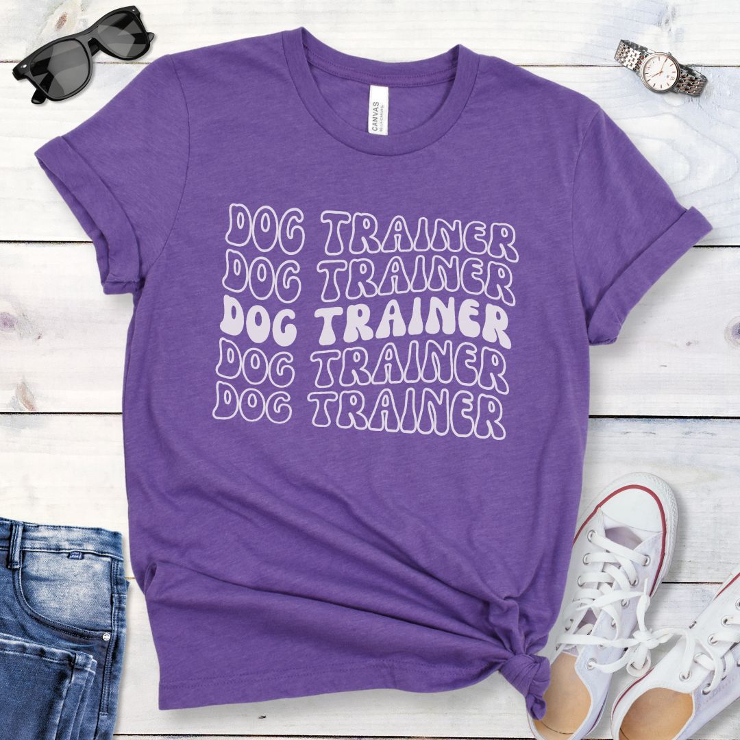 Purple dog training t-shirt with 'DOG TRAINER' text in repeated, groovy retro font, presented on a wooden surface with casual items around.