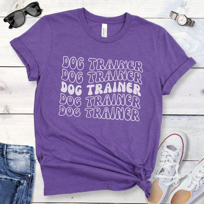 Purple dog training t-shirt with 'DOG TRAINER' text in repeated, groovy retro font, presented on a wooden surface with casual items around.