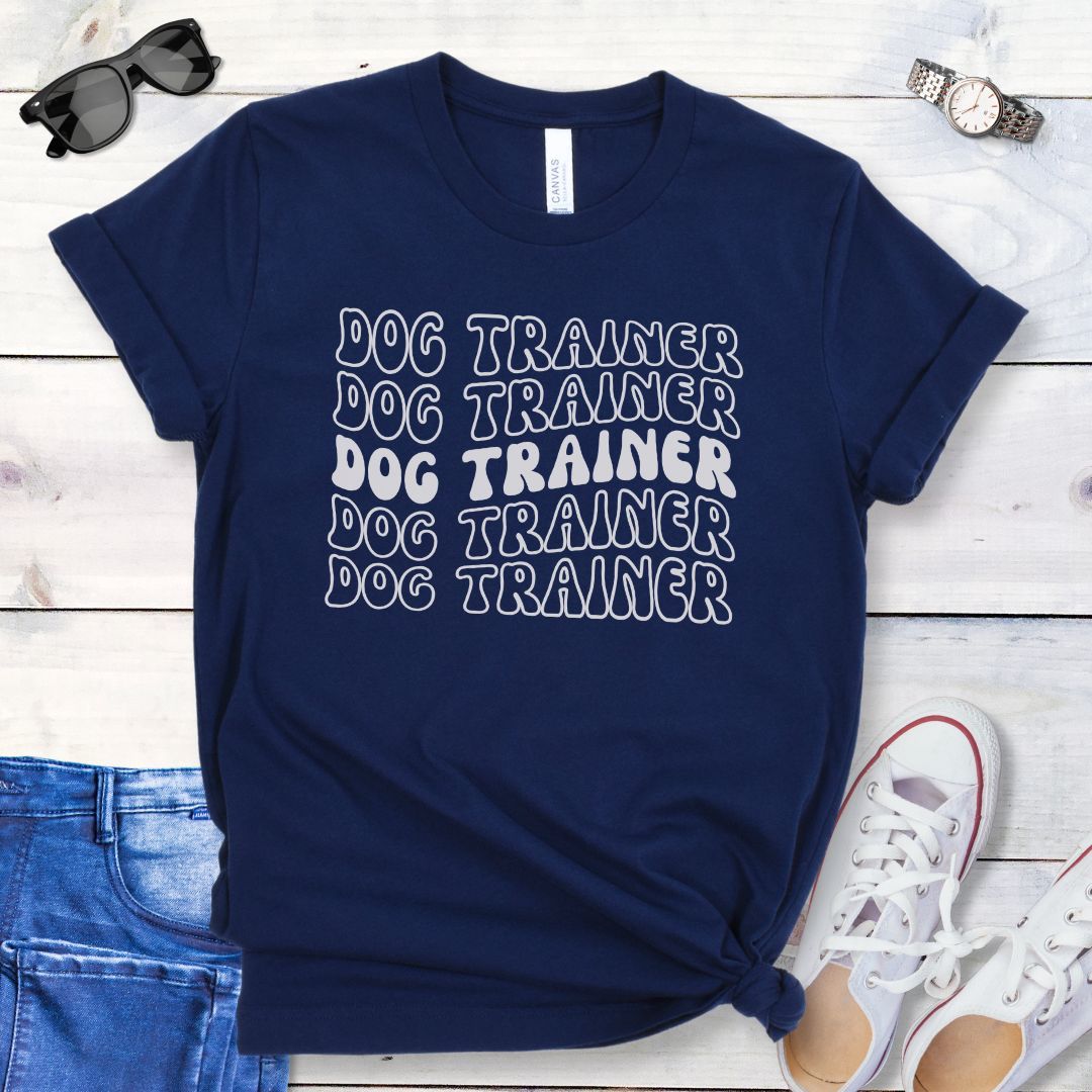 Navy blue dog training t-shirt with 'DOG TRAINER' text in repeated, groovy retro font, presented on a wooden surface with casual items around.