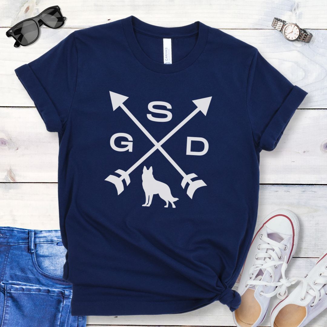 Navy blue t-shirt with a graphic of crossed arrows, the letters GSD, and silhouette of a German Shepherd Dog in the center, displayed on a wooden surface with a pair of jeans and white shoes in the background.