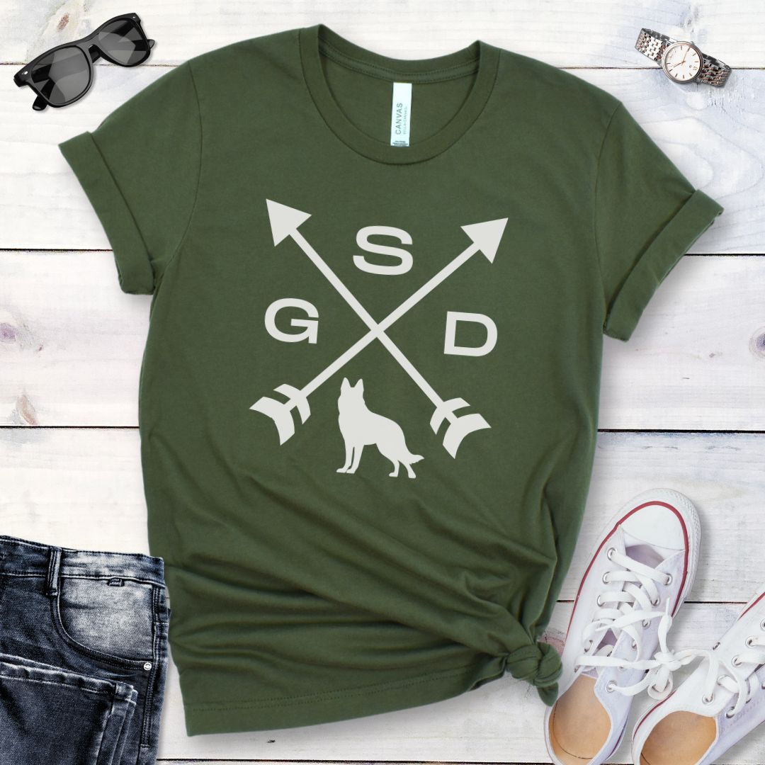 Military green t-shirt with a graphic of crossed arrows, the letters GSD, and silhouette of a German Shepherd Dog in the center, displayed on a wooden surface with a pair of jeans and white shoes in the background.