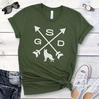 Military green t-shirt with a graphic of crossed arrows, the letters GSD, and silhouette of a German Shepherd Dog in the center, displayed on a wooden surface with a pair of jeans and white shoes in the background.