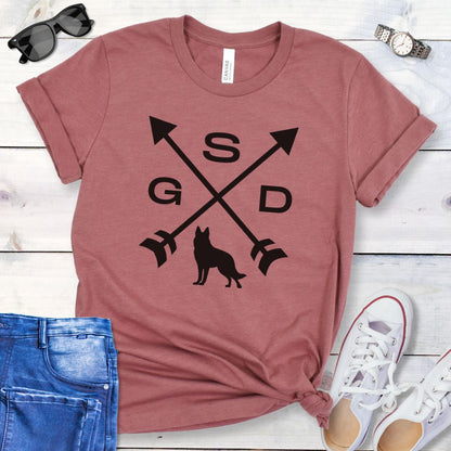 Mauve t-shirt with a graphic of crossed arrows, the letters GSD, and silhouette of a German Shepherd Dog in the center, displayed on a wooden surface with a pair of jeans and white shoes in the background.