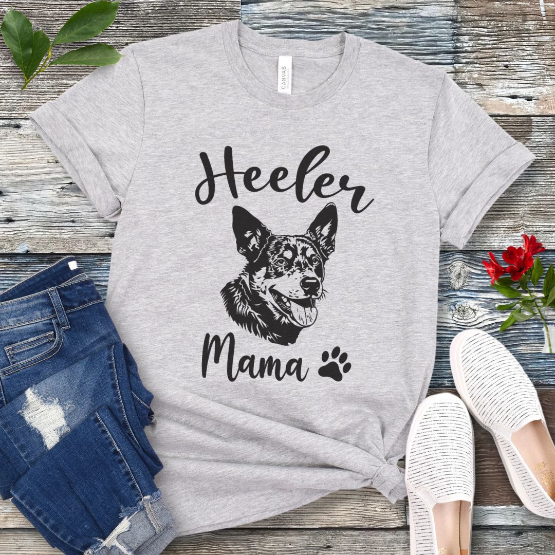 A grey dog lover T-shirt with 'Heeler Mama' text in script font and a graphic of a smiling Australian Cattle Dog face, accompanied by a paw print. The shirt is displayed on a wooden surface with a pair of jeans and white shoes in the background.