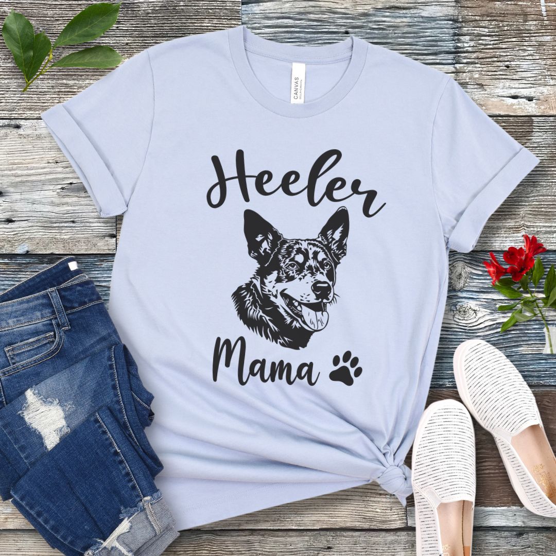 A light blue dog lover T-shirt with 'Heeler Mama' text in script font and a graphic of a smiling Australian Cattle Dog face, accompanied by a paw print. The shirt is displayed on a wooden surface with a pair of jeans and white shoes in the background.