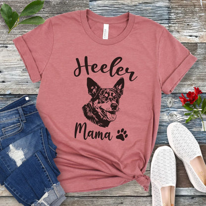 A mauve dog lover T-shirt with 'Heeler Mama' text in script font and a graphic of a smiling Australian Cattle Dog face, accompanied by a paw print. The shirt is displayed on a wooden surface with a pair of jeans and white shoes in the background.