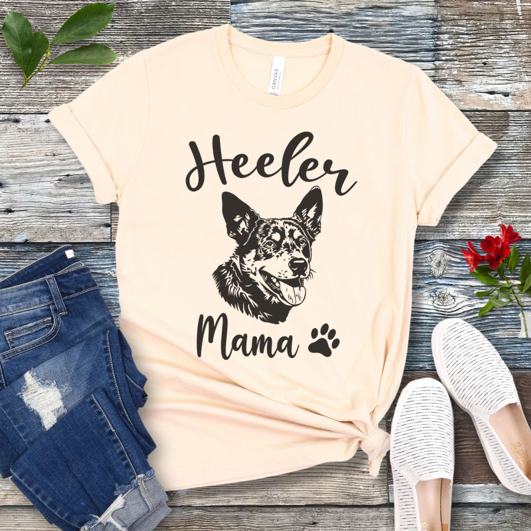 A beige dog lover T-shirt with 'Heeler Mama' text in script font and a graphic of a smiling Australian Cattle Dog face, accompanied by a paw print. The shirt is displayed on a wooden surface with a pair of jeans and white shoes in the background.
