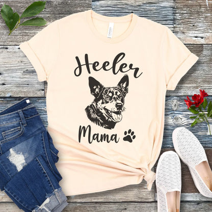 A beige dog lover T-shirt with 'Heeler Mama' text in script font and a graphic of a smiling Australian Cattle Dog face, accompanied by a paw print. The shirt is displayed on a wooden surface with a pair of jeans and white shoes in the background.