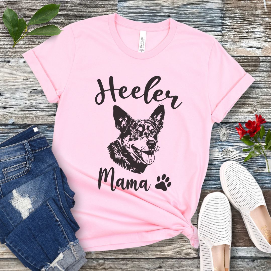 A pink dog lover T-shirt with 'Heeler Mama' text in script font and a graphic of a smiling Australian Cattle Dog face, accompanied by a paw print. The shirt is displayed on a wooden surface with a pair of jeans and white shoes in the background.