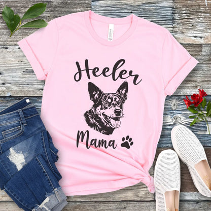 A pink dog lover T-shirt with 'Heeler Mama' text in script font and a graphic of a smiling Australian Cattle Dog face, accompanied by a paw print. The shirt is displayed on a wooden surface with a pair of jeans and white shoes in the background.