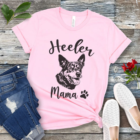 A pink dog lover T-shirt with 'Heeler Mama' text in script font and a graphic of a smiling Australian Cattle Dog face, accompanied by a paw print. The shirt is displayed on a wooden surface with a pair of jeans and white shoes in the background.