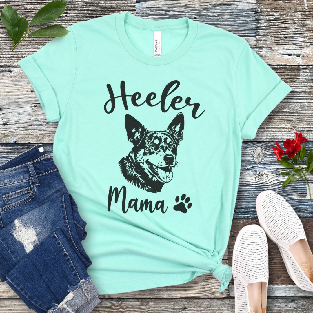 A mint green dog lover T-shirt with 'Heeler Mama' text in script font and a graphic of a smiling Australian Cattle Dog face, accompanied by a paw print. The shirt is displayed on a wooden surface with a pair of jeans and white shoes in the background.