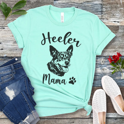 A mint green dog lover T-shirt with 'Heeler Mama' text in script font and a graphic of a smiling Australian Cattle Dog face, accompanied by a paw print. The shirt is displayed on a wooden surface with a pair of jeans and white shoes in the background.