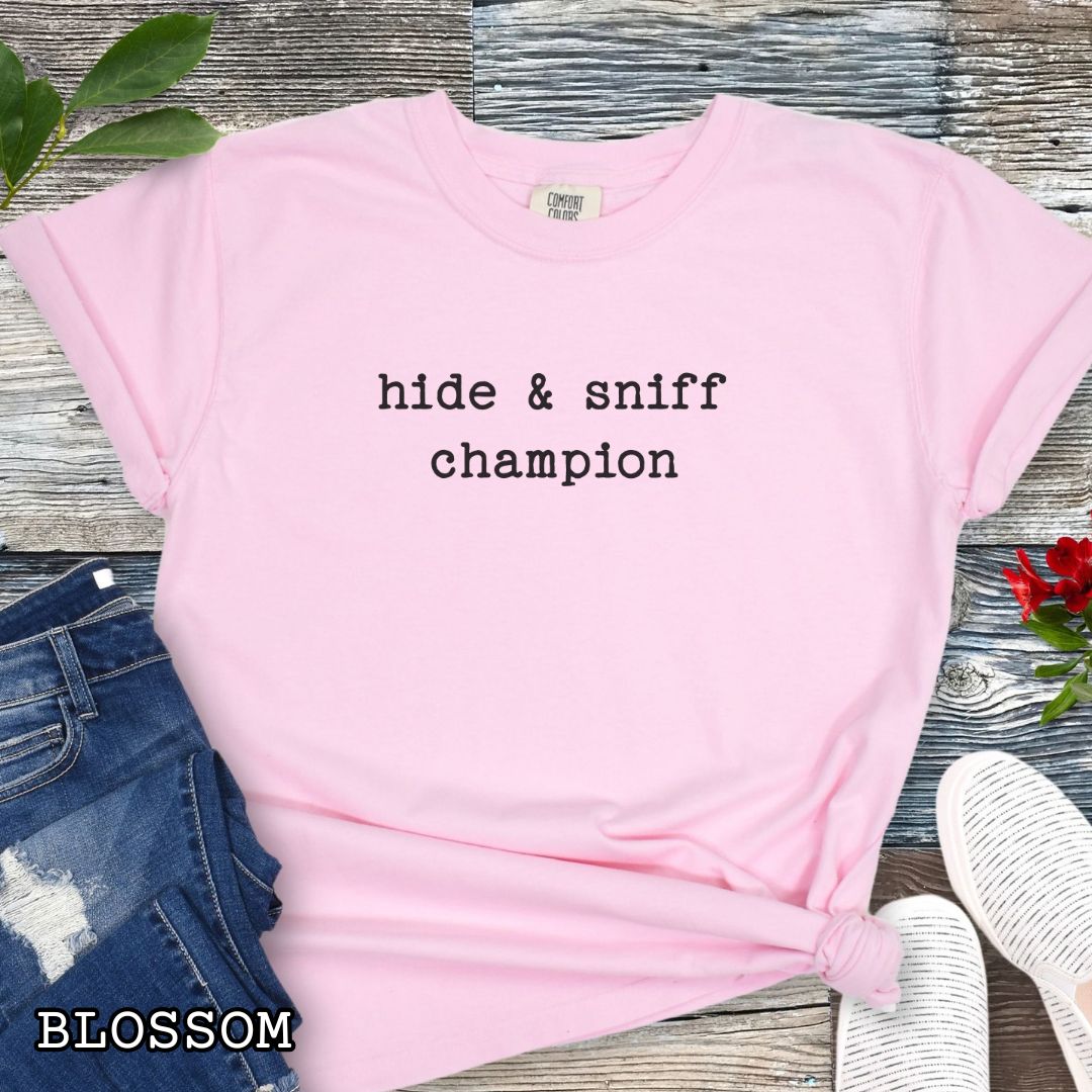 Pink Comfort Colors dog scent training t-shirt with 'hide & sniff champion' text, paired with denim shorts and a red flower on a wooden surface.
