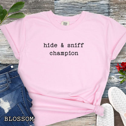 Pink Comfort Colors dog scent training t-shirt with 'hide & sniff champion' text, paired with denim shorts and a red flower on a wooden surface.