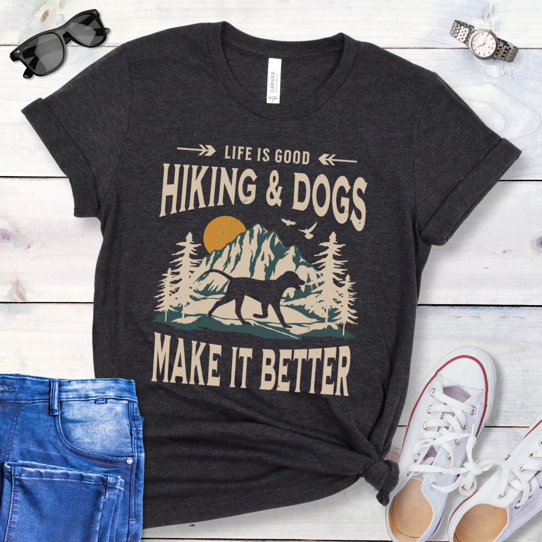 Dark grey t-shirt for outdoorsy dog lovers with text 'LIFE IS GOOD HIKING & DOGS MAKE IT BETTER' and a vintage style illustration of mountains,  pine trees and sunset with a trotting dog, paired with blue jeans and white sneakers.