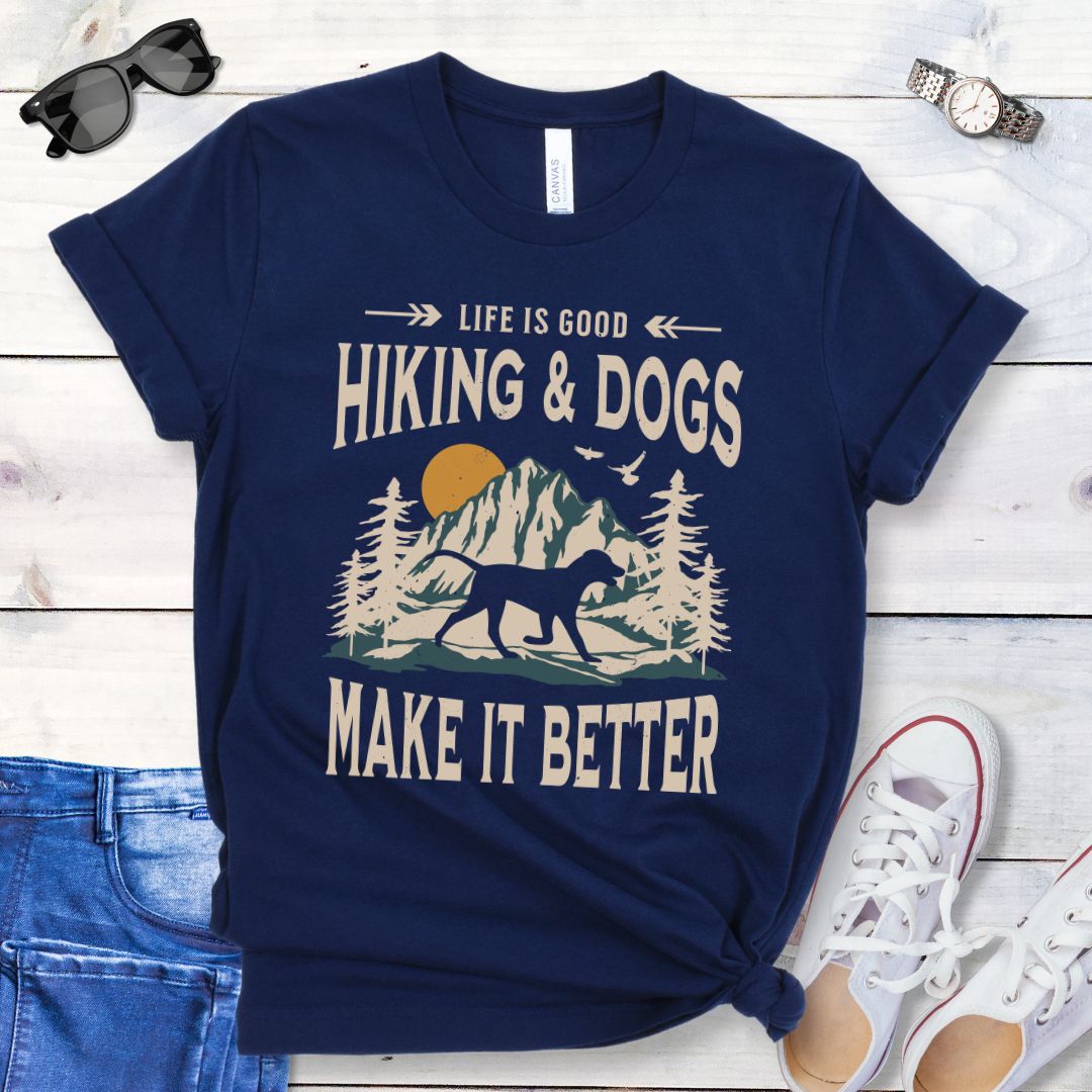Navy t-shirt for outdoorsy dog lovers with text 'LIFE IS GOOD HIKING & DOGS MAKE IT BETTER' and a vintage style illustration of mountains,  pine trees and sunset with a trotting dog, paired with blue jeans and white sneakers.