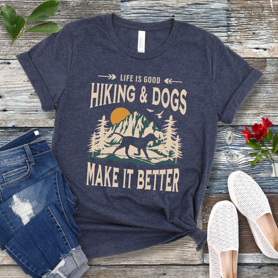 Heather navy t-shirt for outdoorsy dog lovers with text 'LIFE IS GOOD HIKING & DOGS MAKE IT BETTER' and a vintage style illustration of mountains,  pine trees and sunset with a trotting dog, paired with blue jeans and white sneakers.