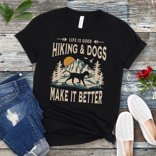 Black t-shirt for outdoorsy dog lovers with text 'LIFE IS GOOD HIKING & DOGS MAKE IT BETTER' and a vintage style illustration of mountains,  pine trees and sunset with a trotting dog, paired with blue jeans and white sneakers.