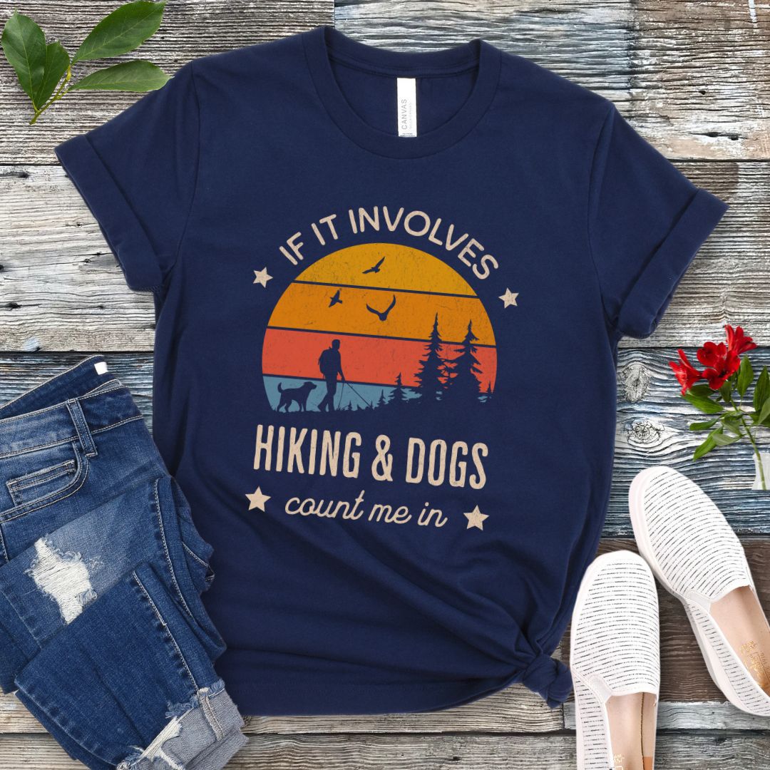 Navy blue T-shirt with text 'IF IT INVOLVES HIKING & DOGS COUNT ME IN' printed on the front, featuring a sunset scene and hiker and dog silhouettes. The shirt is laid out flat with a pair of blue jeans and white sneakers.