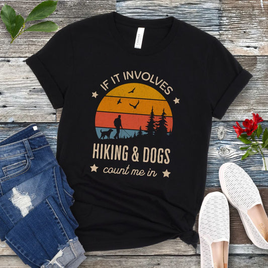 Black T-shirt with text 'IF IT INVOLVES HIKING & DOGS COUNT ME IN' printed on the front, featuring a sunset scene and hiker and dog silhouettes. The shirt is laid out flat with a pair of blue jeans and white sneakers.