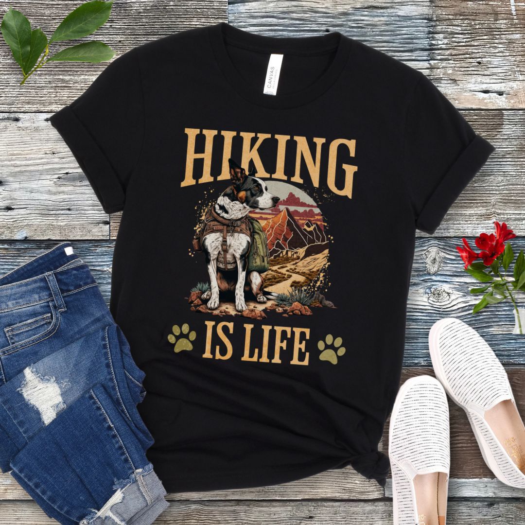 Black t-shirt with an image of a dog on a mountain trial and text 'HIKING IS LIFE', displayed on a wooden surface with a pair of jeans and white shoes in the background.