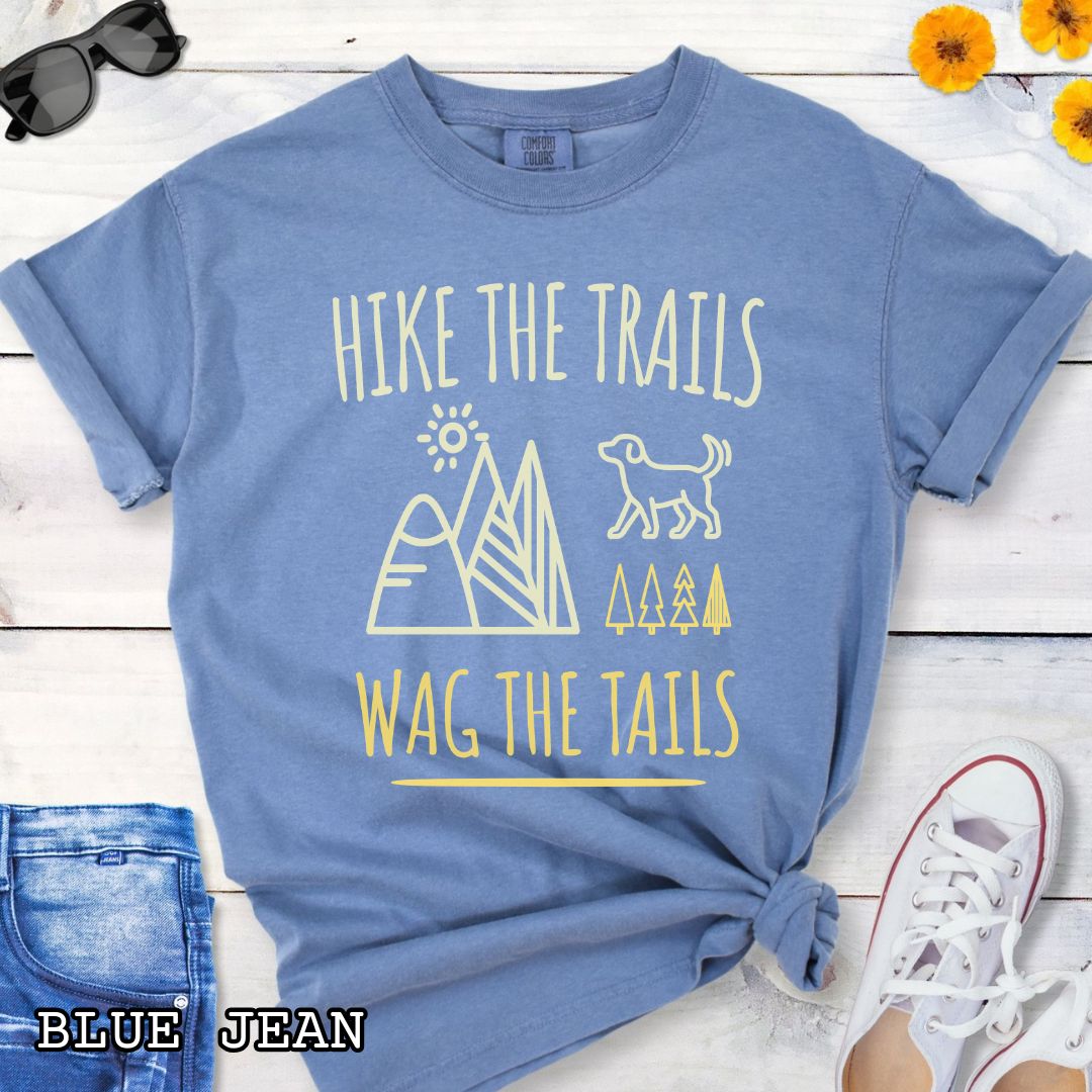 Blue Comfort Colors t-shirt for dog-loving hikers and campers with playful minimalist 'Hike the Trails, Wag the Tails' design, shown on a wooden surface with jeans and sneakers.