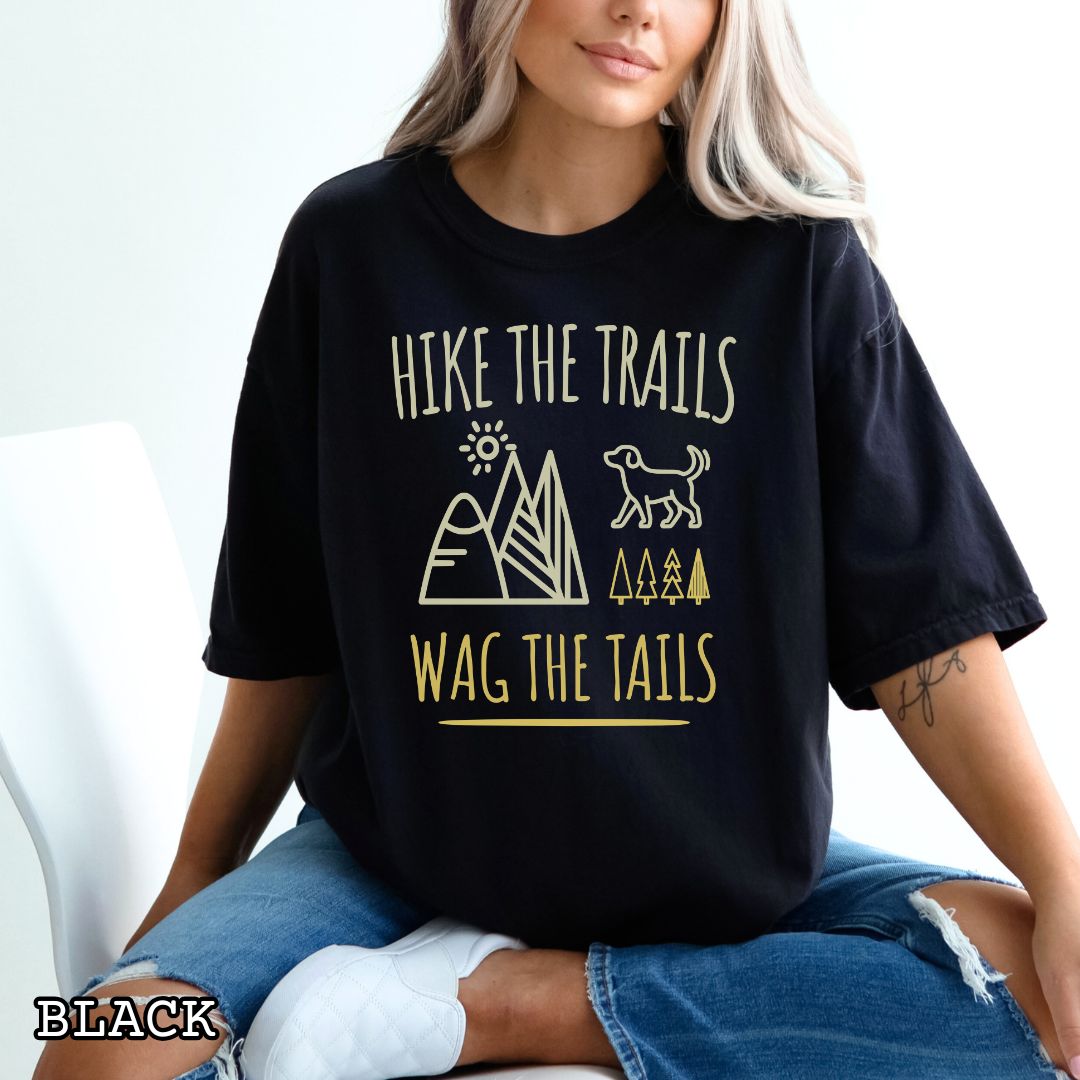 Black Comfort Colors t-shirt for dog-loving hikers and campers with playful minimalist 'Hike the Trails, Wag the Tails' design, shown on a wooden surface with jeans and sneakers.