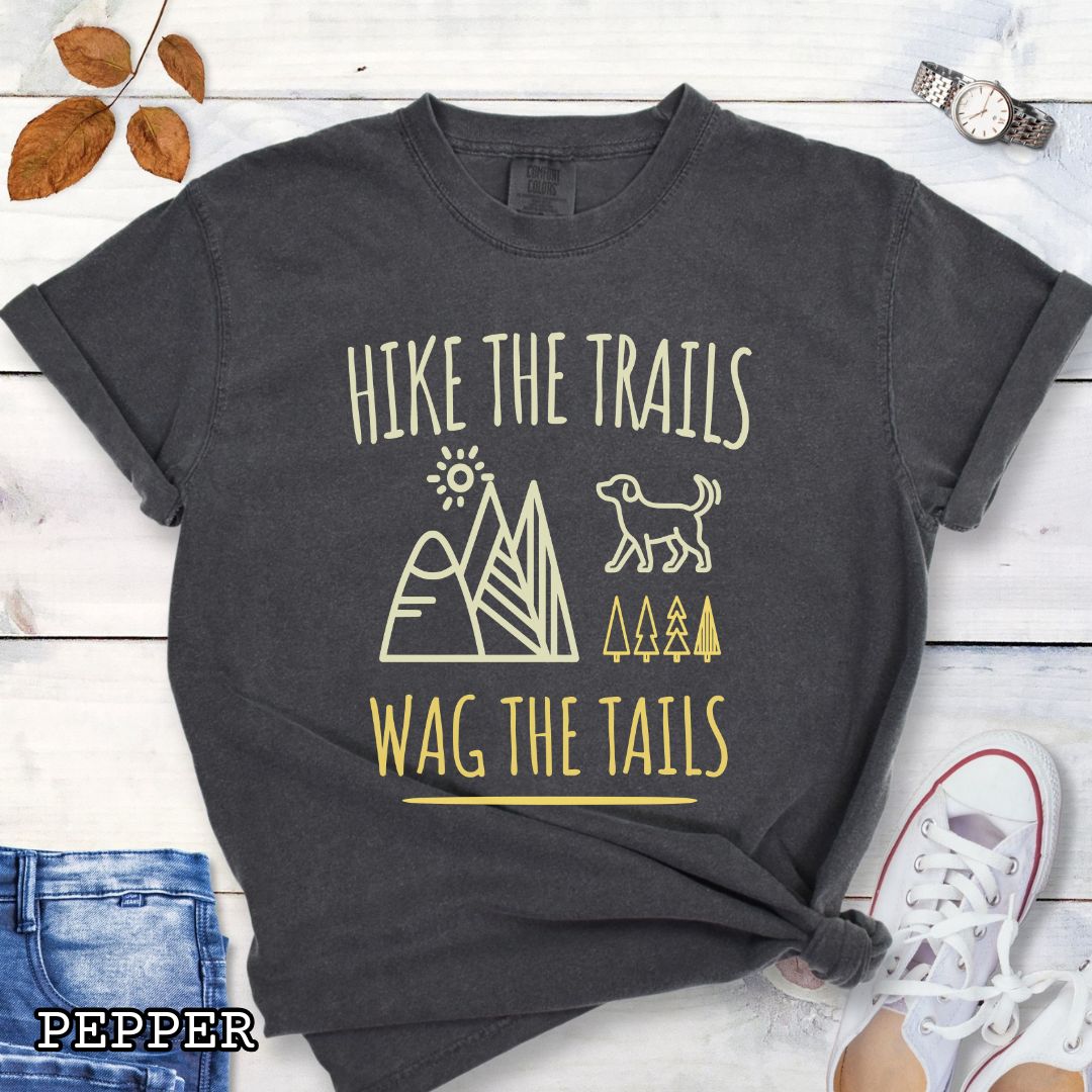 Dark grey Comfort Colors t-shirt for dog-loving hikers and campers with playful minimalist 'Hike the Trails, Wag the Tails' design, shown on a wooden surface with jeans and sneakers.