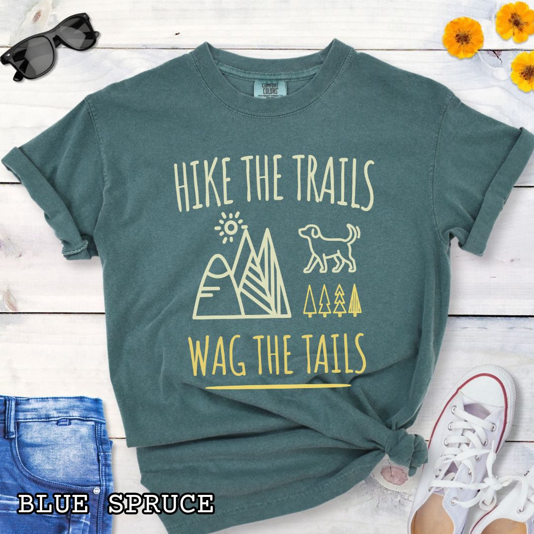 Green Comfort Colors t-shirt for dog-loving hikers and campers with playful minimalist 'Hike the Trails, Wag the Tails' design, shown on a wooden surface with jeans and sneakers.