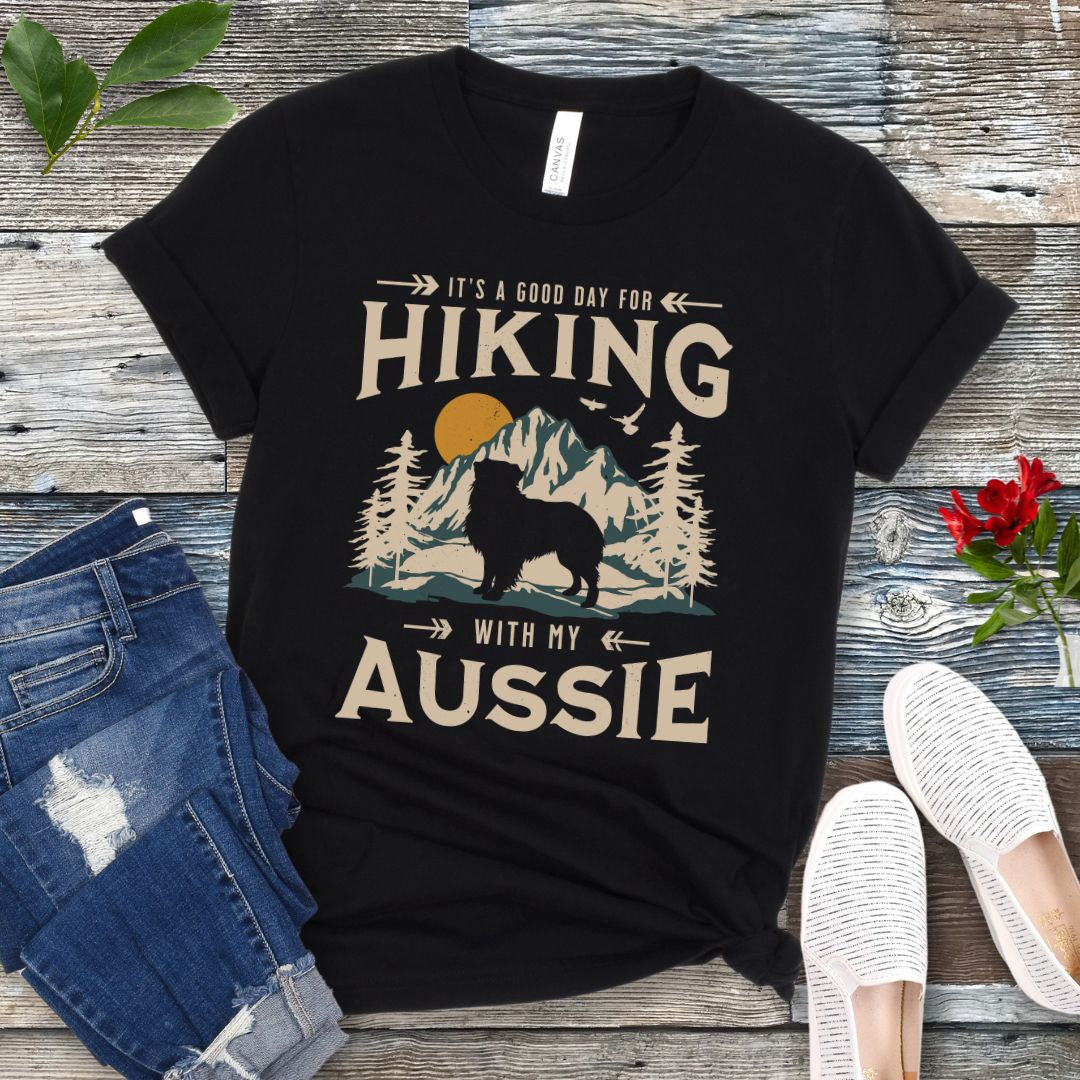 A black t-shirt with a vintage-style graphic print of mountains, an Australian Shepherd silhouette, and the text 'Hiking with my Aussie'. The shirt is presented on a wooden surface with a pair of blue jeans and white shoes in the background.