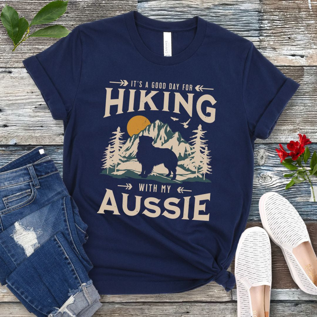 A navy t-shirt with a vintage-style graphic print of mountains, an Australian Shepherd silhouette, and the text 'Hiking with my Aussie'. The shirt is presented on a wooden surface with a pair of blue jeans and white shoes in the background.